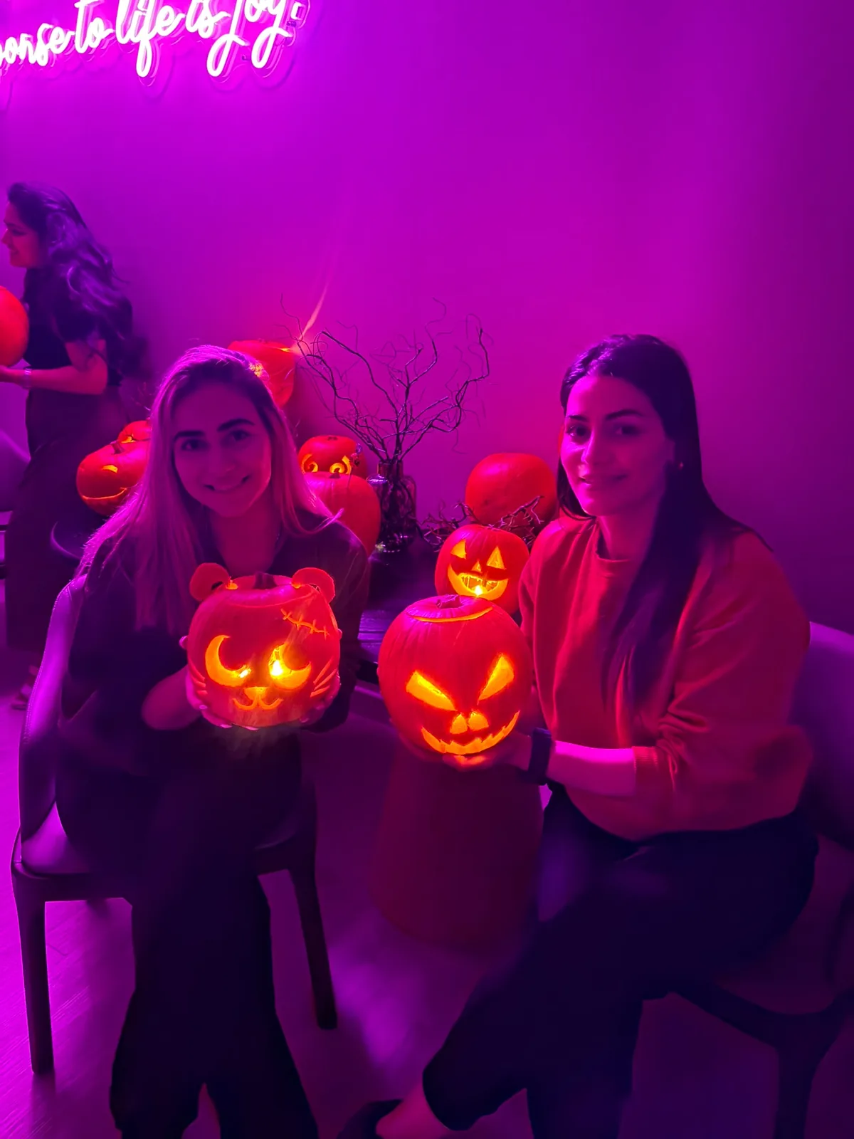 Two residents smiling as they hold up their lit, carved pumpkins in the purple-lit communal lounge