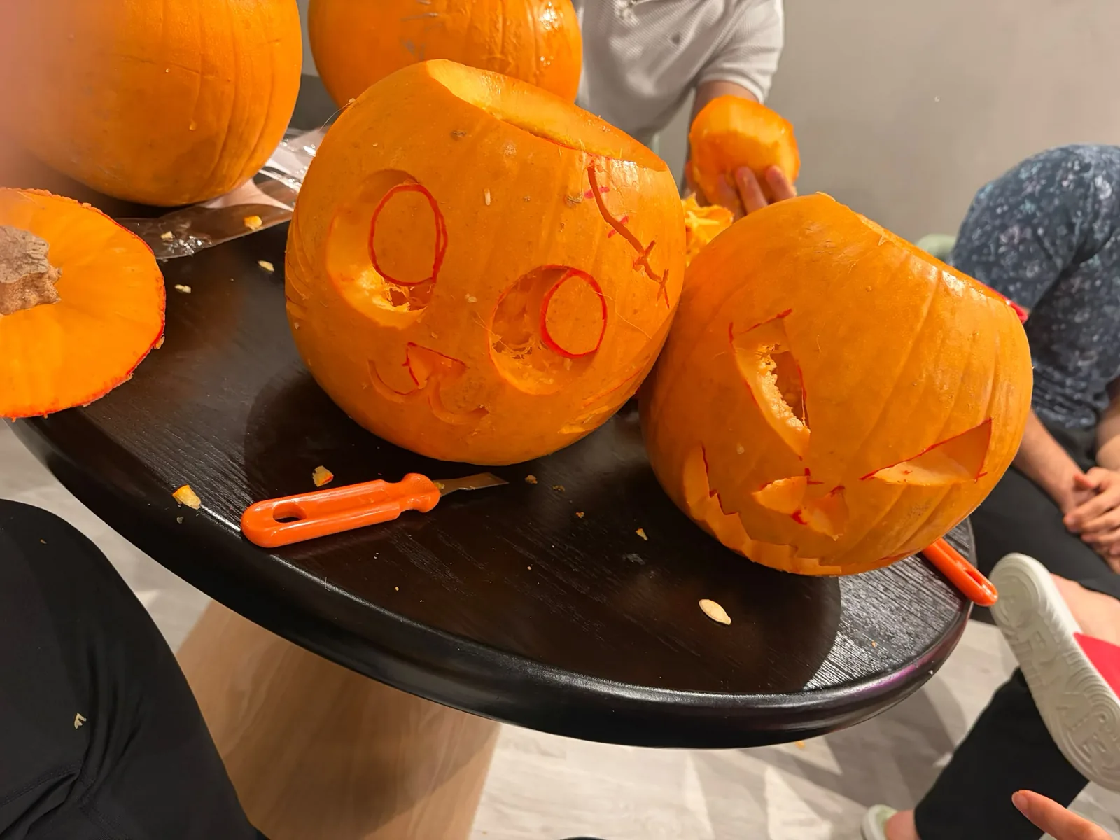 Pumpkins being carved on a round table, with a cat-faced pumpkin, an open pumpkin and orange-handled carving tools