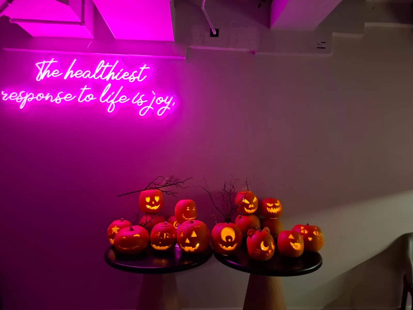 Wide view of the communal lounge with a display of lit pumpkins beneath a pink neon sign reading 'The healthiest response to life is joy'