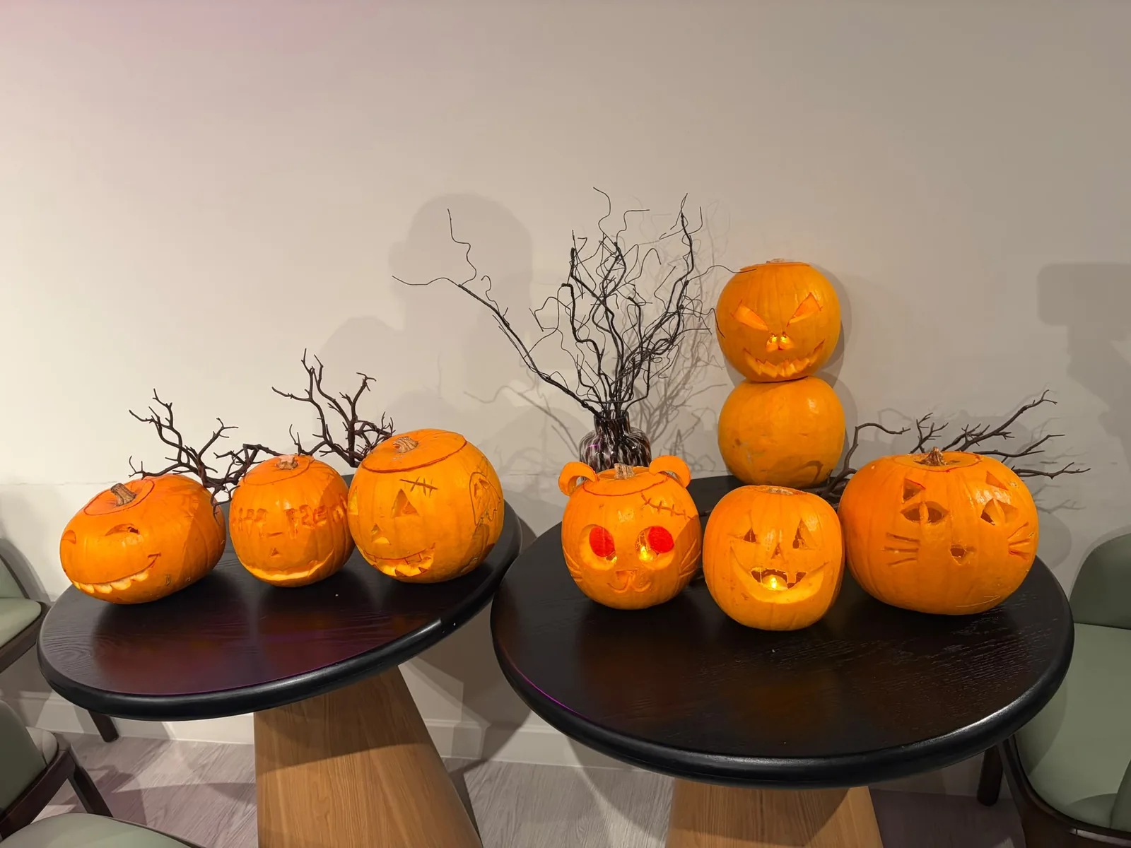 Finished carved pumpkins, including cat faces, a bear-eared pumpkin and a stacked pumpkin tower, arranged on two tables with bare twig decorations